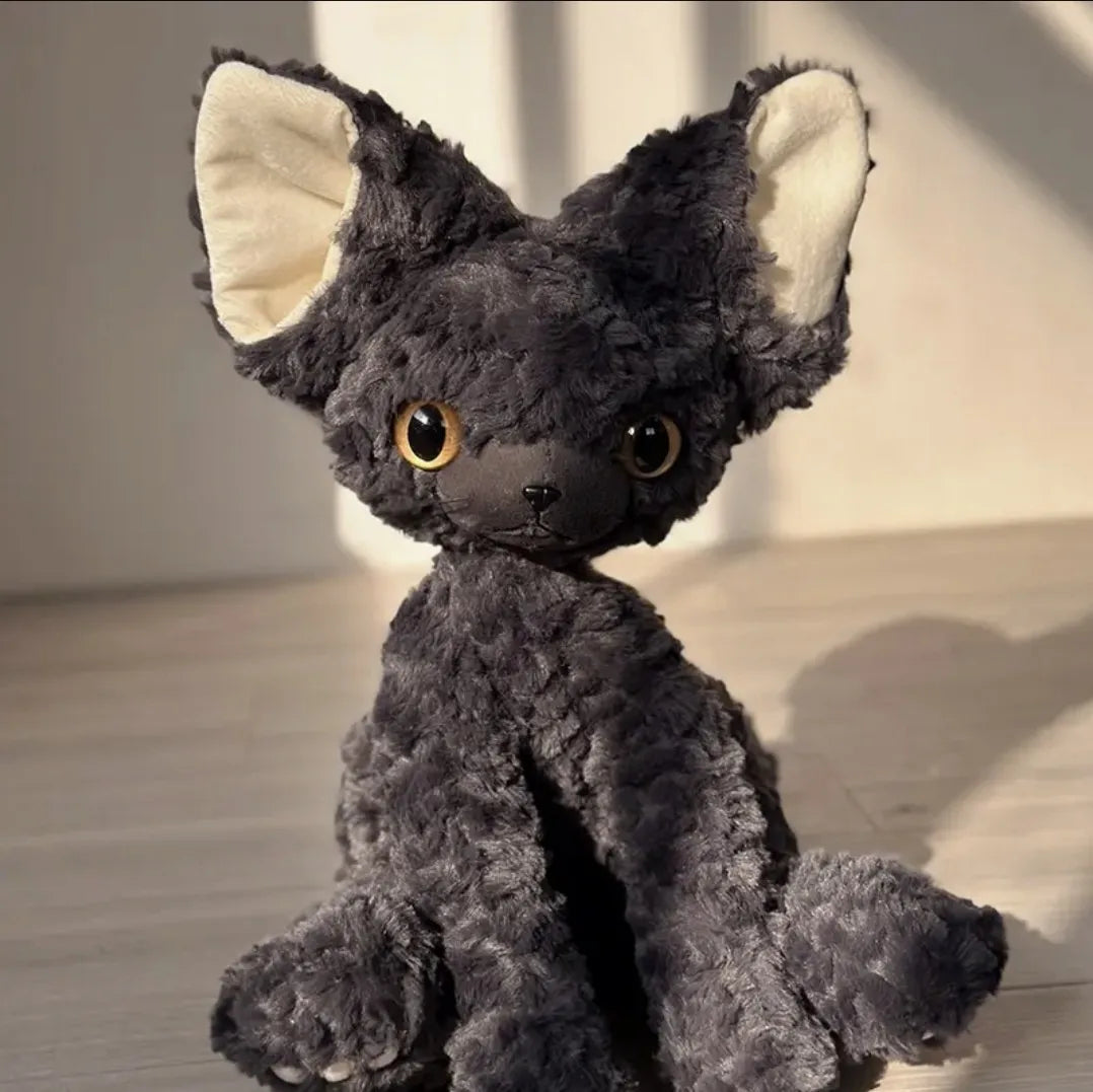 A dark gray curly-haired cat plush toy with cream-colored inner ears and large amber eyes, sitting on a light wood floor in warm natural light.