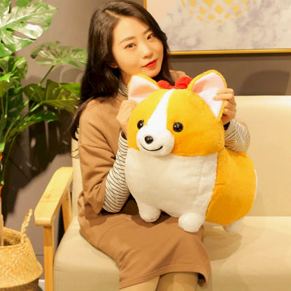 A woman sitting on a beige sofa holds a plush Corgi doll with yellow and white fur, pink inner ears, and a small red bow on its head.