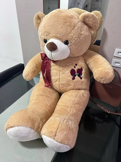 Large beige teddy bear with a red bow and heart design on a chair.