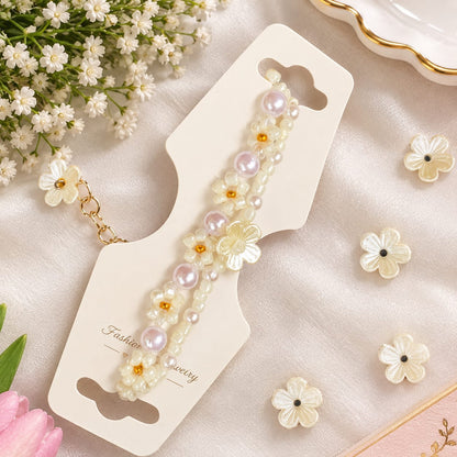 A beaded floral bracelet displayed on a cream card, surrounded by small white flower charms, baby's breath, and soft pink petals on a satin fabric background.