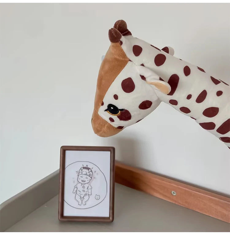 Plush giraf toy leaning against a white wall with a framed illustration of a baby on a shelf.