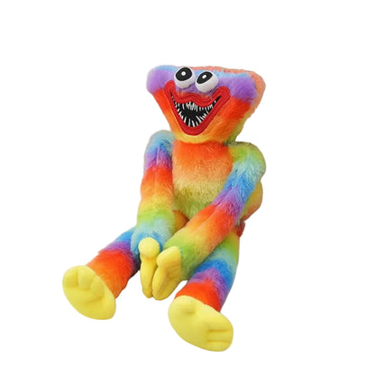 A plush toy resembling a rainbow monster with bright multicolor fur, large round eyes, a wide toothy grin, and yellow feet, posed on a light fabric surface beside a woven basket.