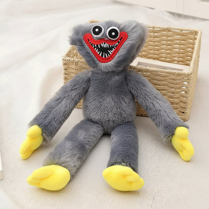 A plush toy resembling a gray monster with large round eyes, a wide red grin showing white and black teeth, and bright yellow feet, posed on a light fabric surface beside a woven basket.