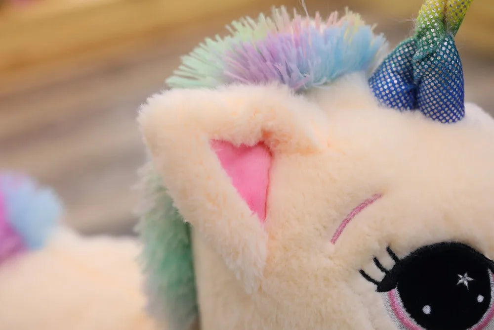 Close-up of a plush unicorn toy with cream-colored fur, pastel rainbow mane, pink inner ears, embroidered eyelashes, and a blue glittery horn bow.