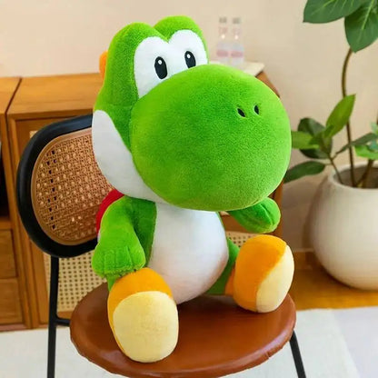 A green plush dinosaur yoshi plush toy with white and orange accents, styled sitting on a brown chair in a cozy indoor room with a potted plant in the background.